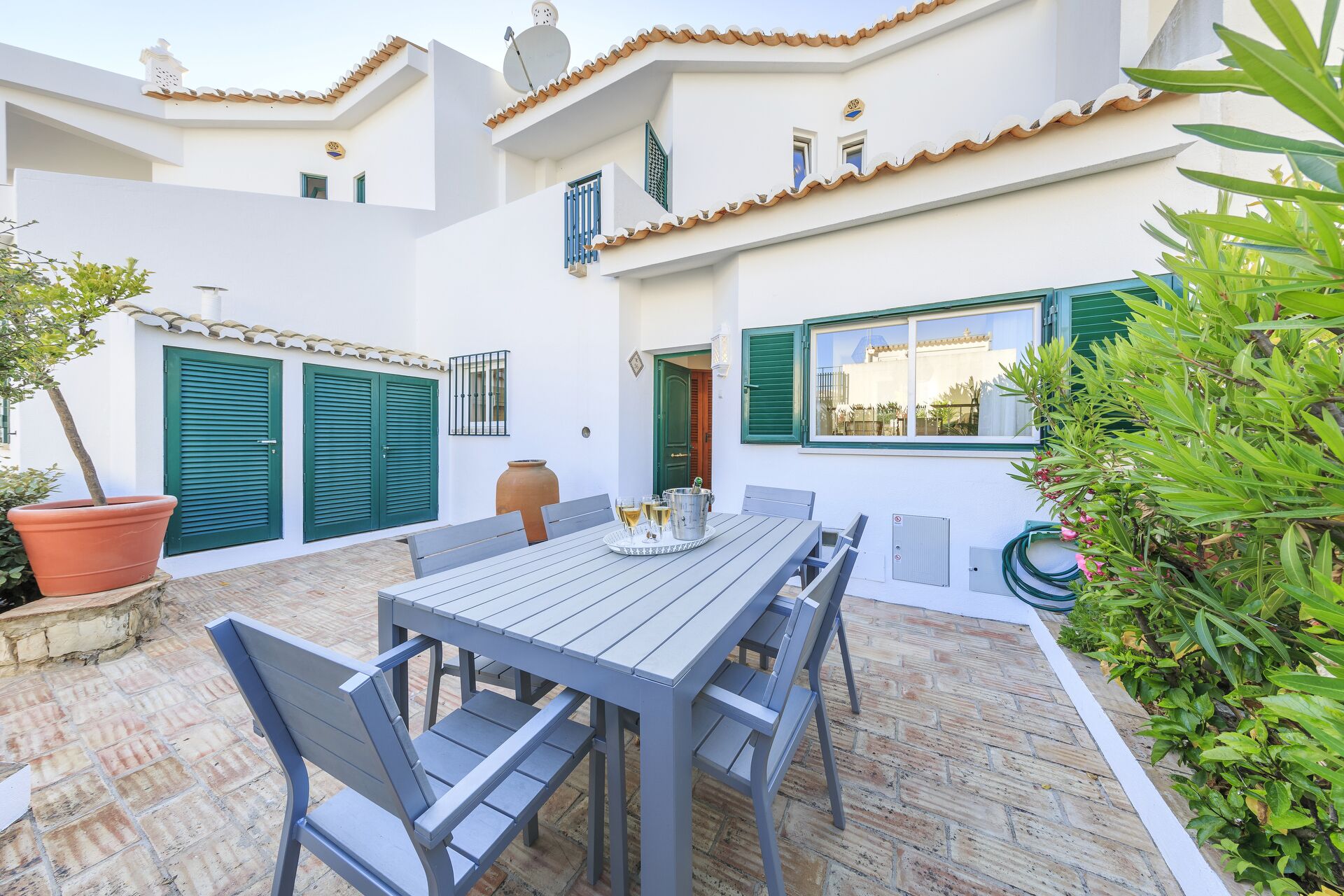 A patio with a table and chairs, a large potted plant, and a view of a building with green shutters and a tiled roof.