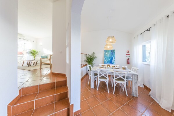 A dining area with a table set for four, white chairs, and a hanging light fixture. - CASAS DA PRAIA, 56 (Almancil)