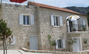 Un bâtiment en pierre avec des volets blancs, un balcon doté d’une rambarde en métal et une montagne en arrière-plan.