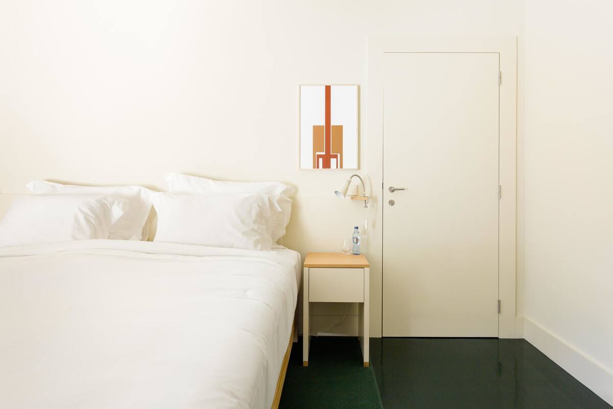 A hotel room with a bed, a bedside table with a bottle, and a door.