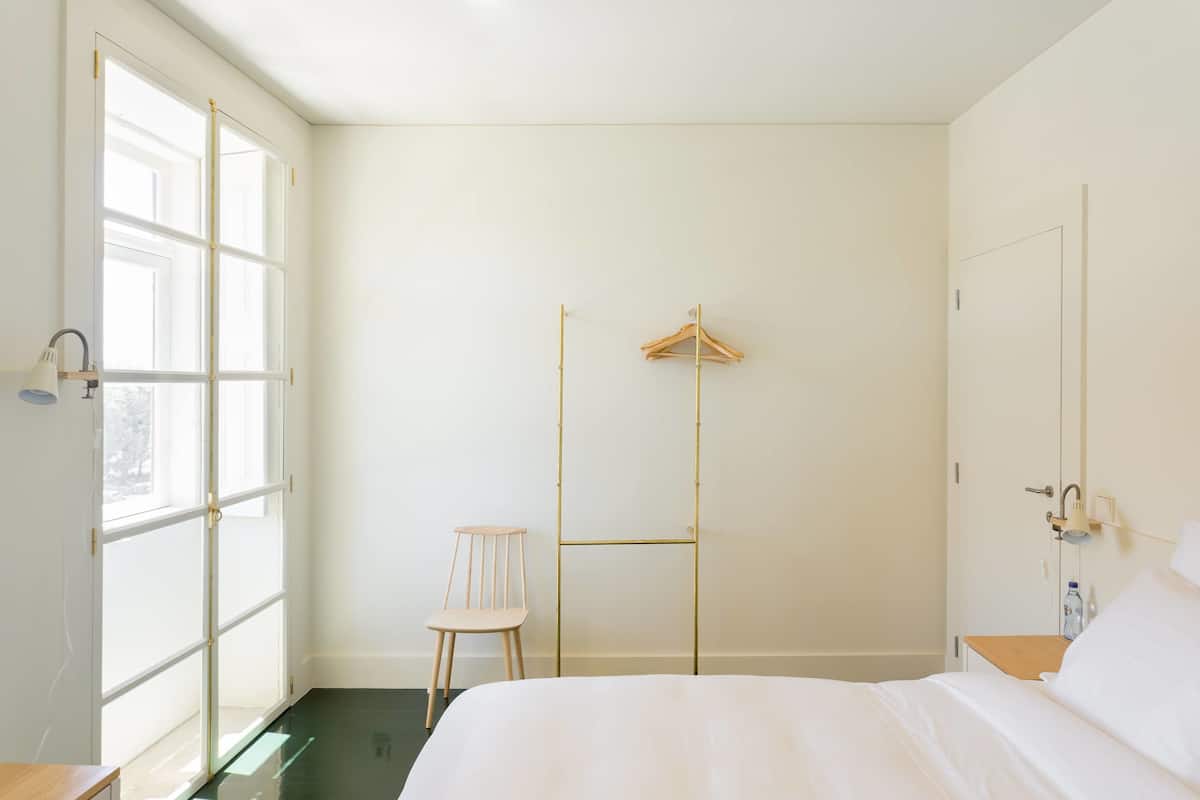 A hotel room with a bed, a chair, a wooden hanger, and a window.