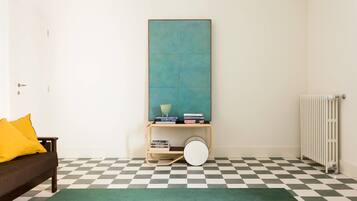 Una habitación con suelo de cuadros, una alfombra verde, un sofá con cojines amarillos, una estantería con libros y un cuenco, y un radiador.