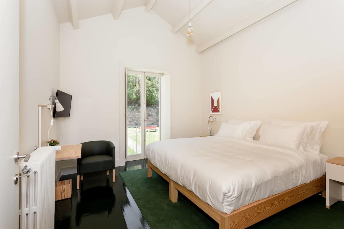 A bedroom with a large bed, a desk with a chair, a radiator, and a window with a view of greenery.