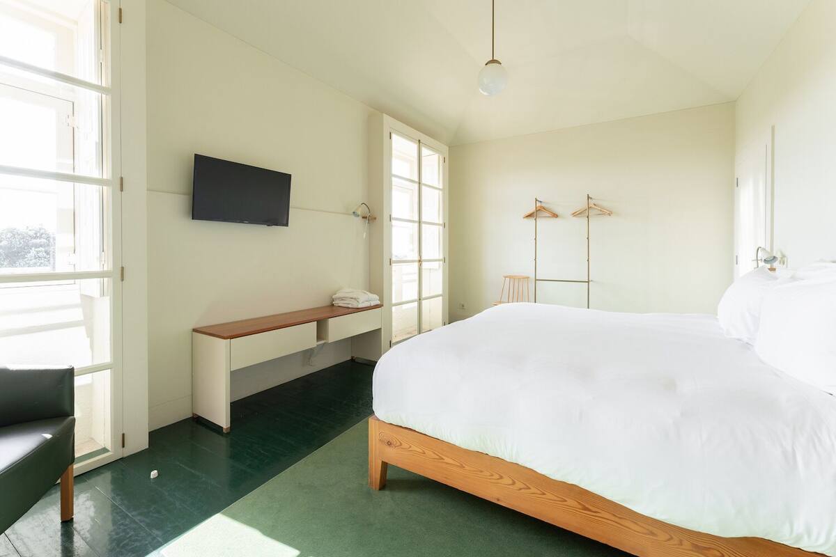 A modern hotel room with a large bed, a TV mounted on the wall, a bench with a towel, and a clothing rack.