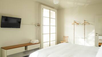 A hotel room with a bed, a TV mounted on the wall, a bench with towels, a chair, and a wardrobe with hangers.