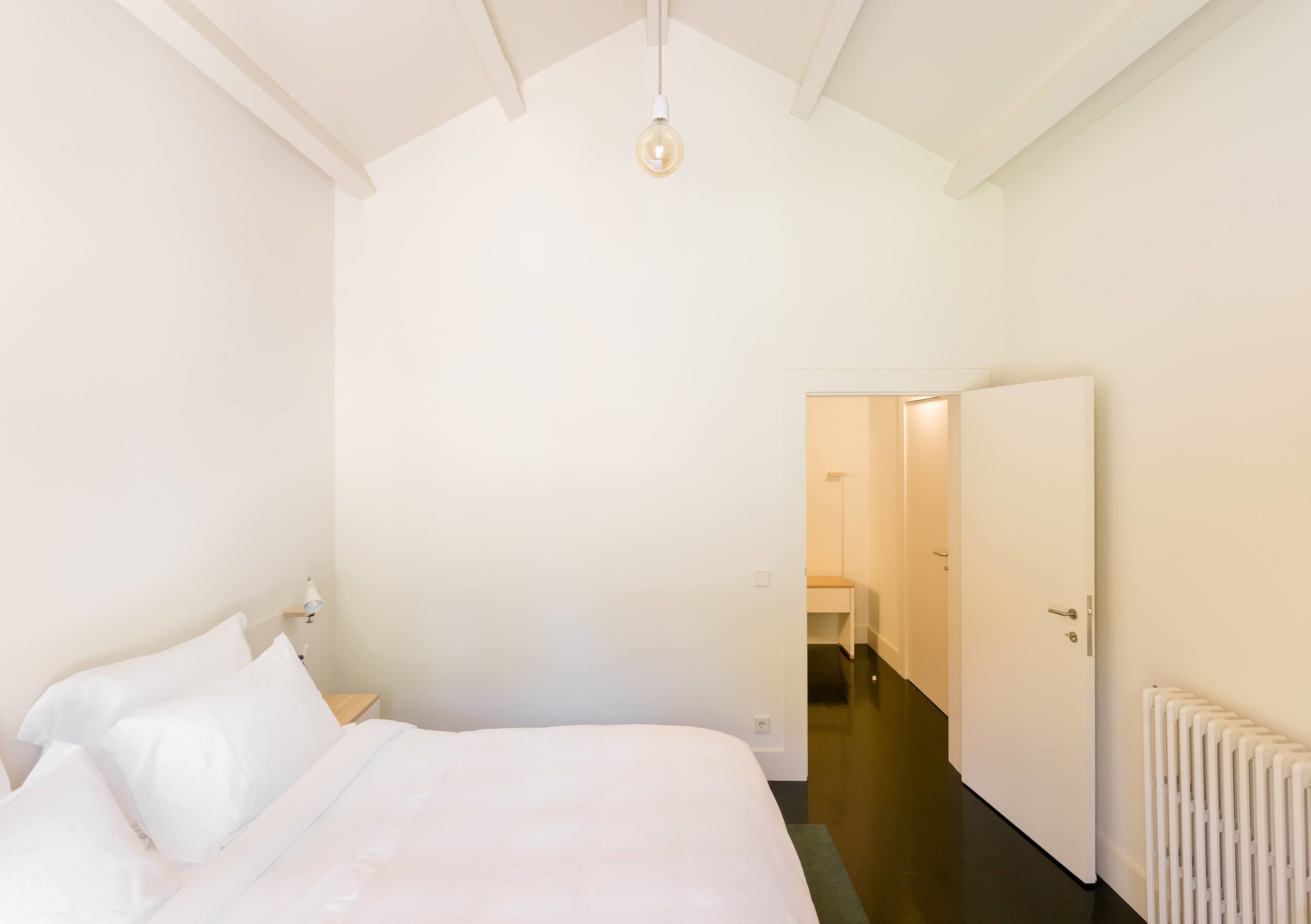 A bedroom with a bed, a bedside table, a hanging light, and a door leading to another room.