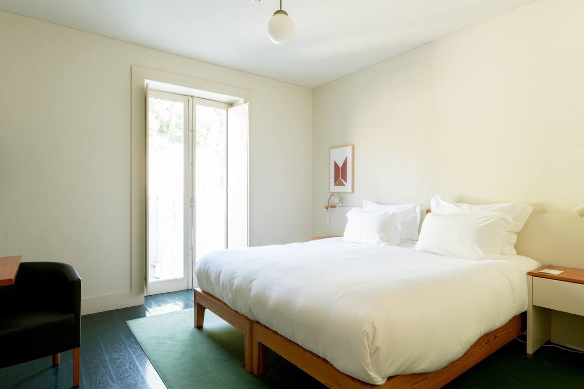 A bedroom with a large bed, a desk, a chair, and a window with a view of greenery.