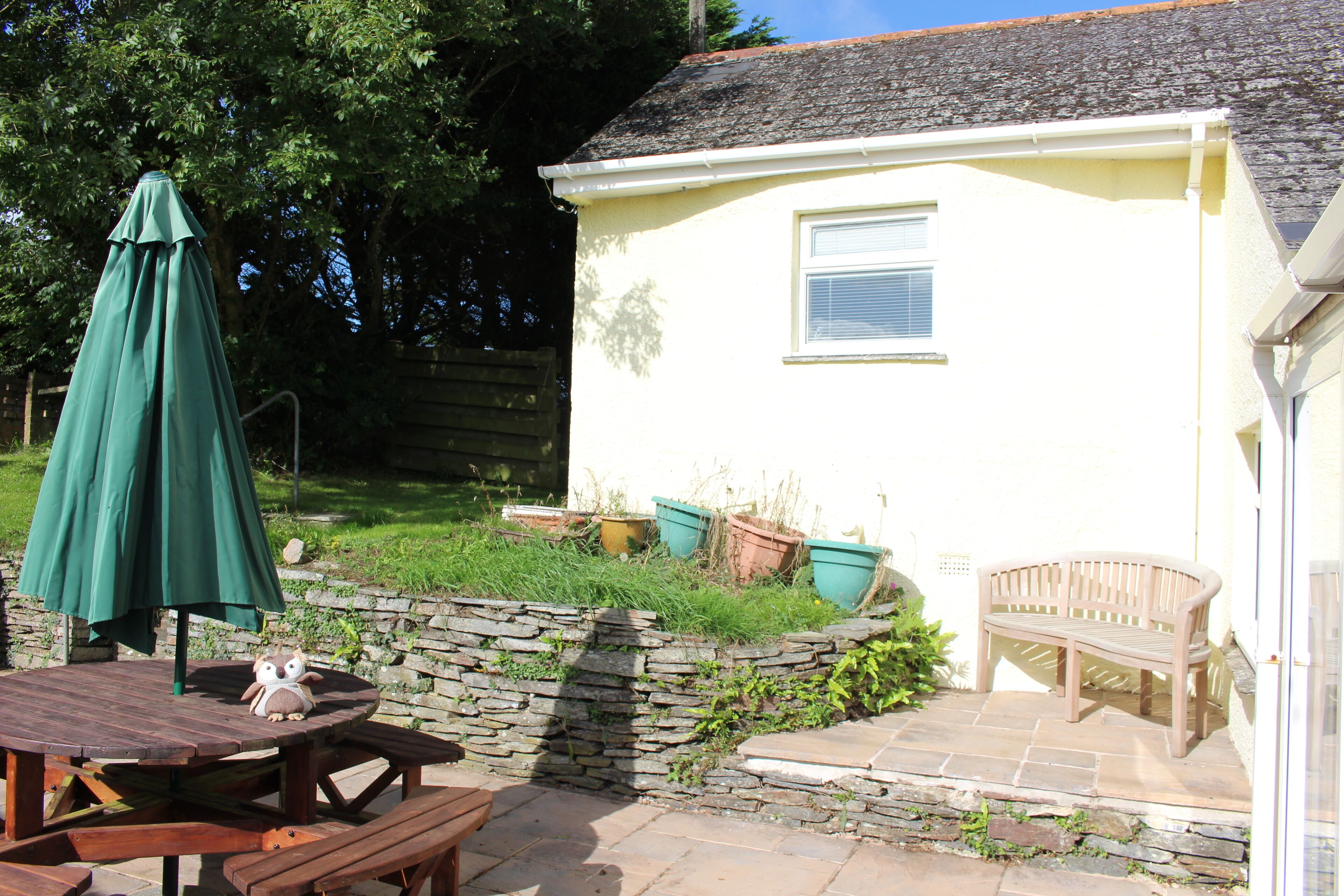A patio area with a wooden table, a green umbrella, a stone wall, and a bench.