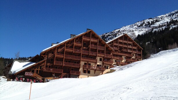 Ein Chalet-Stil Gebäude aus Holz mit mehreren Balkonen, gelegen an einem verschneiten Hang unter blauem Himmel.