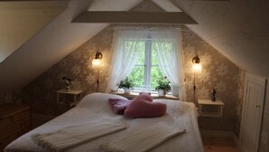 A bedroom with a sloped ceiling, a bed with white linens, two bedside tables with lamps, a window with sheer curtains, and a small potted plant on the windowsill. - Nice old style cottage close to the Baltic Sea (Gryt, Valdemarsvik)