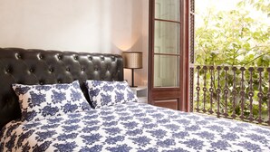 Une chambre à coucher avec une tête de lit capitonnée, une literie à motifs floraux, une lampe de chevet et une vue sur la verdure par la fenêtre.