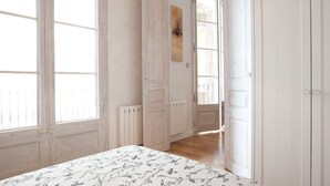 Une pièce dotée de grandes fenêtres, d’un radiateur, d’un lit avec une literie à motifs floraux et d’une porte donnant sur une autre pièce.