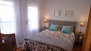 Ein ordentlich eingerichtetes Schlafzimmer mit einem Bett, zwei Nachttischlampen, einem Sessel und einem Fenster mit durchsichtigen Vorhängen.