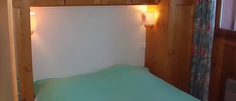 A bedroom with a green bedspread, wooden cabinets above the bed, and two wall-mounted lamps.
