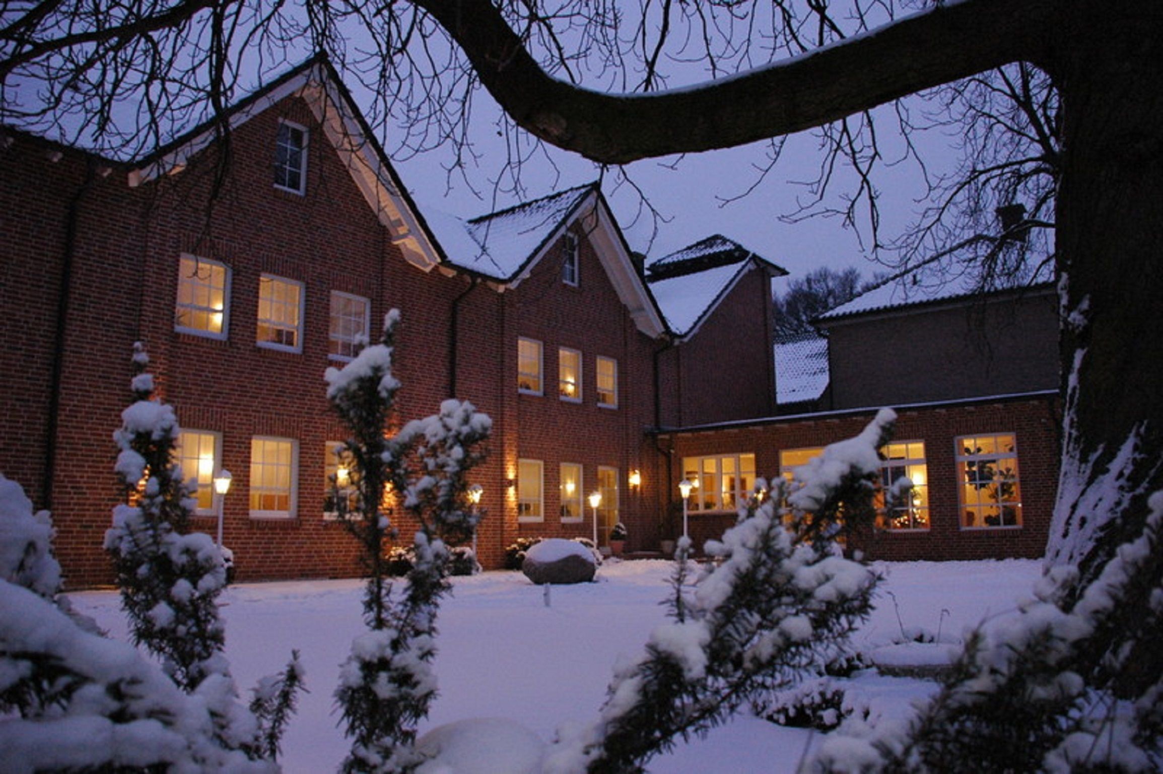 A brick house with multiple windows, a snow-covered yard, and a large tree in the foreground.