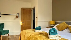 A hotel room with a bed, a green chair, a wooden door, a yellow lamp, and a towel rack.