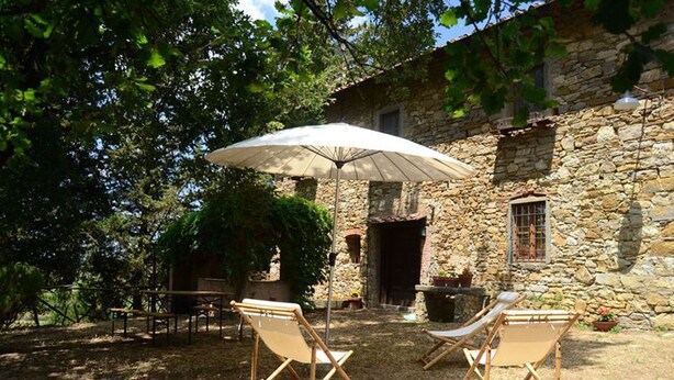 An outdoor seating area with two wooden chairs and a large umbrella, set against a stone building with a rustic appearance. - Ancient stone house in the heart of the Chianti Classico (Panzano in Chianti)