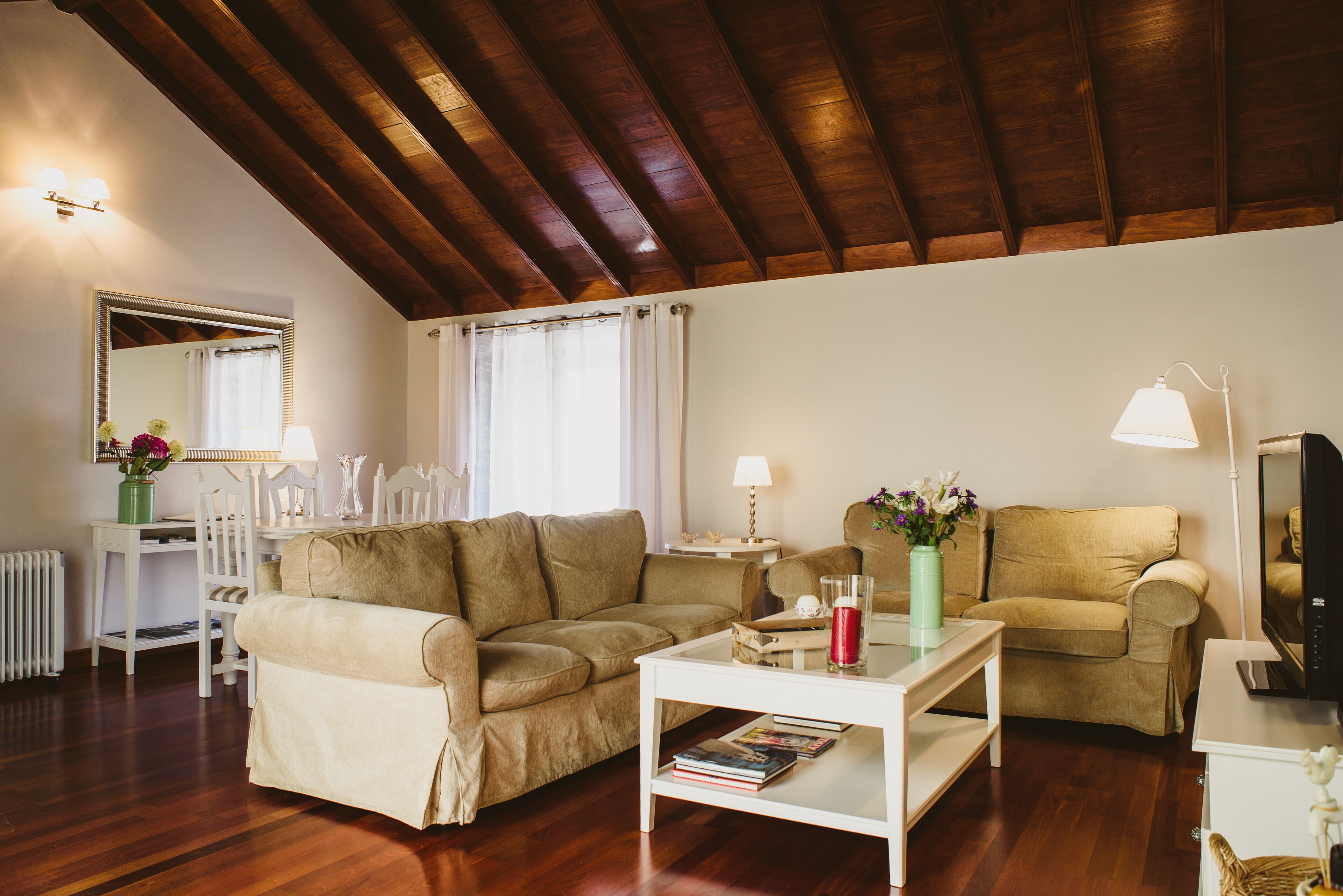 A cozy living room with a wooden ceiling, two sofas, a coffee table with books, a TV, and a fireplace.