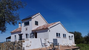 Una casa de dos pisos con pared de piedra, techo de tejas y un pequeño patio con mobiliario exterior.