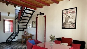 Una habitación con piso a cuadros, una escalera, una mesa con mantel rojo, un sofá y un cartel en la pared.