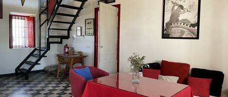 Una habitación con piso a cuadros, una escalera, una mesa con mantel rojo, un sofá y un cartel en la pared.