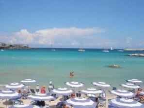 A beach with rows of sun umbrellas and lounge chairs, a clear blue sea, and boats in the distance. - Otranto: Apartment/ flat - OtrantoLuxury Apartment (Otranto)