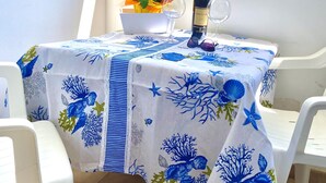 Ein Balkon mit Tisch, gedeckt für zwei Personen. Die blaue und weiße Tischdecke zeigt Muster von Korallen und Meereslebewesen. Auf dem Tisch stehen eine Weinflasche und zwei Gläser, zusammen mit einem gelben Servietten und einem Blumenstrauß.