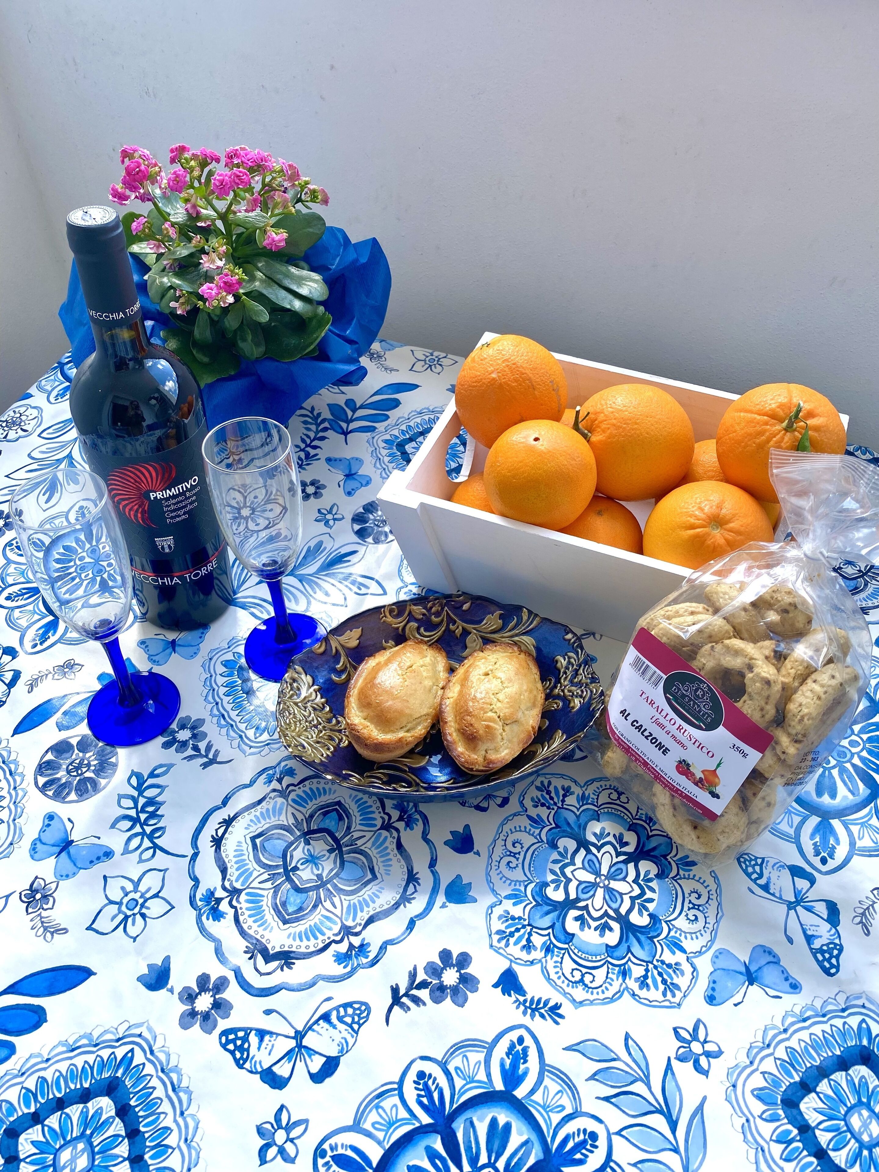 Une table avec une bouteille de vin, une boîte de mandarines, une assiette de biscuits et un vase rempli de fleurs.