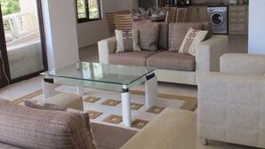 Ein modernes Wohnzimmer mit einem Glastisch, beigen Sofas und einem Essbereich mit Holzsesseln. Im Hintergrund ist eine Waschmaschine zu sehen.
