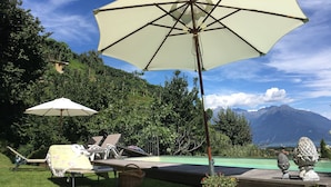 Zona junto a la piscina con sillas reclinables y sombrillas, rodeada de abundante vegetación y montañas al fondo.