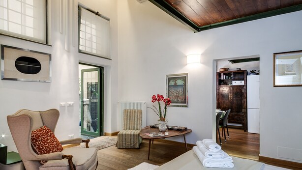 A spacious room with a large bed, a chair, a table with a vase of flowers, and a view of a kitchen and dining area through an open door. - NAVONA ROMANTIC LOFT (Roma)