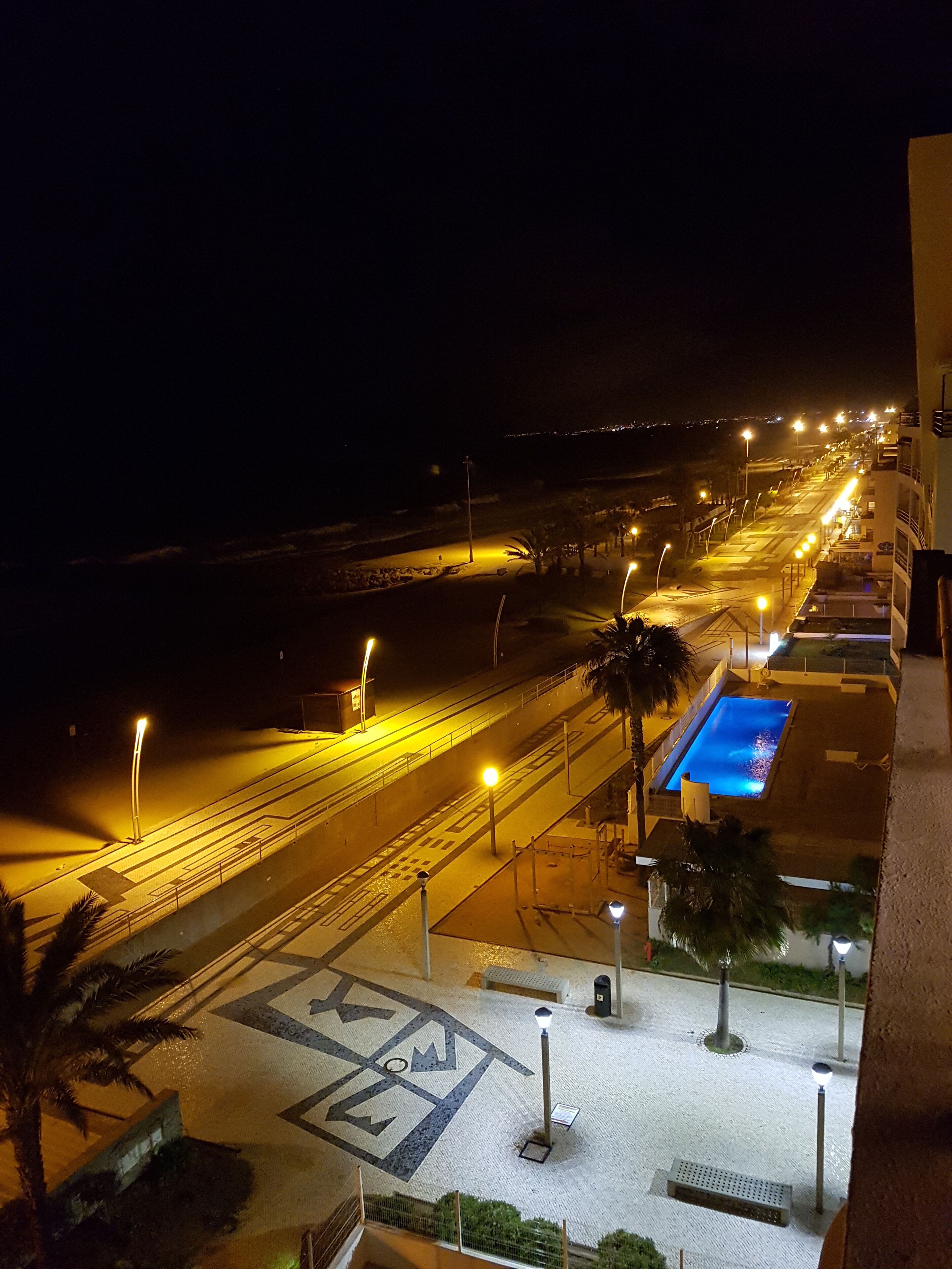 A nighttime view of a well-lit urban area with a pool, streetlights, and a patterned pavement.