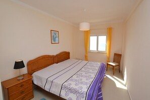 Une chambre à coucher avec un lit, une table de chevet, une chaise, une fenêtre avec des rideaux et un tableau au mur.