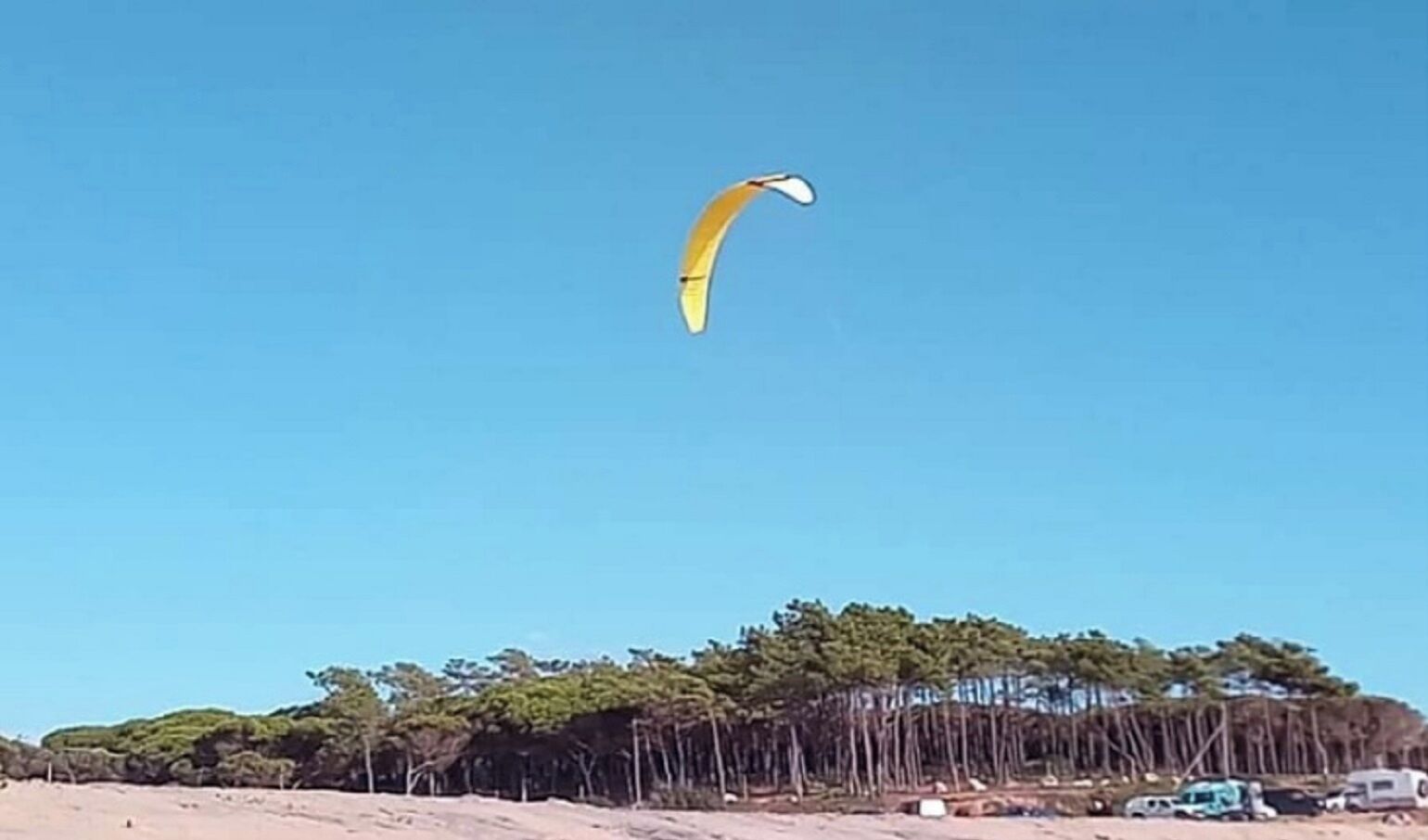 A beach with a clear sky and a yellow parachute-like object. There are trees and a few parked vehicles in the background.