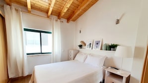 Un dormitorio con techo de madera, una cama con ropa de cama blanca, una mesita de noche con lámpara y una ventana con cortinas.