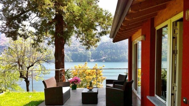 Une terrasse en bois avec des meubles d’extérieur, une vue sur un lac et un bâtiment rouge.