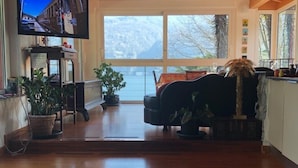 Un vaste salon doté d’un haut plafond en bois, d’une grande fenêtre offrant une vue sur l’extérieur, d’un téléviseur fixé au mur et d’un coin salon confortable agrémenté d’un tapis à motifs.
