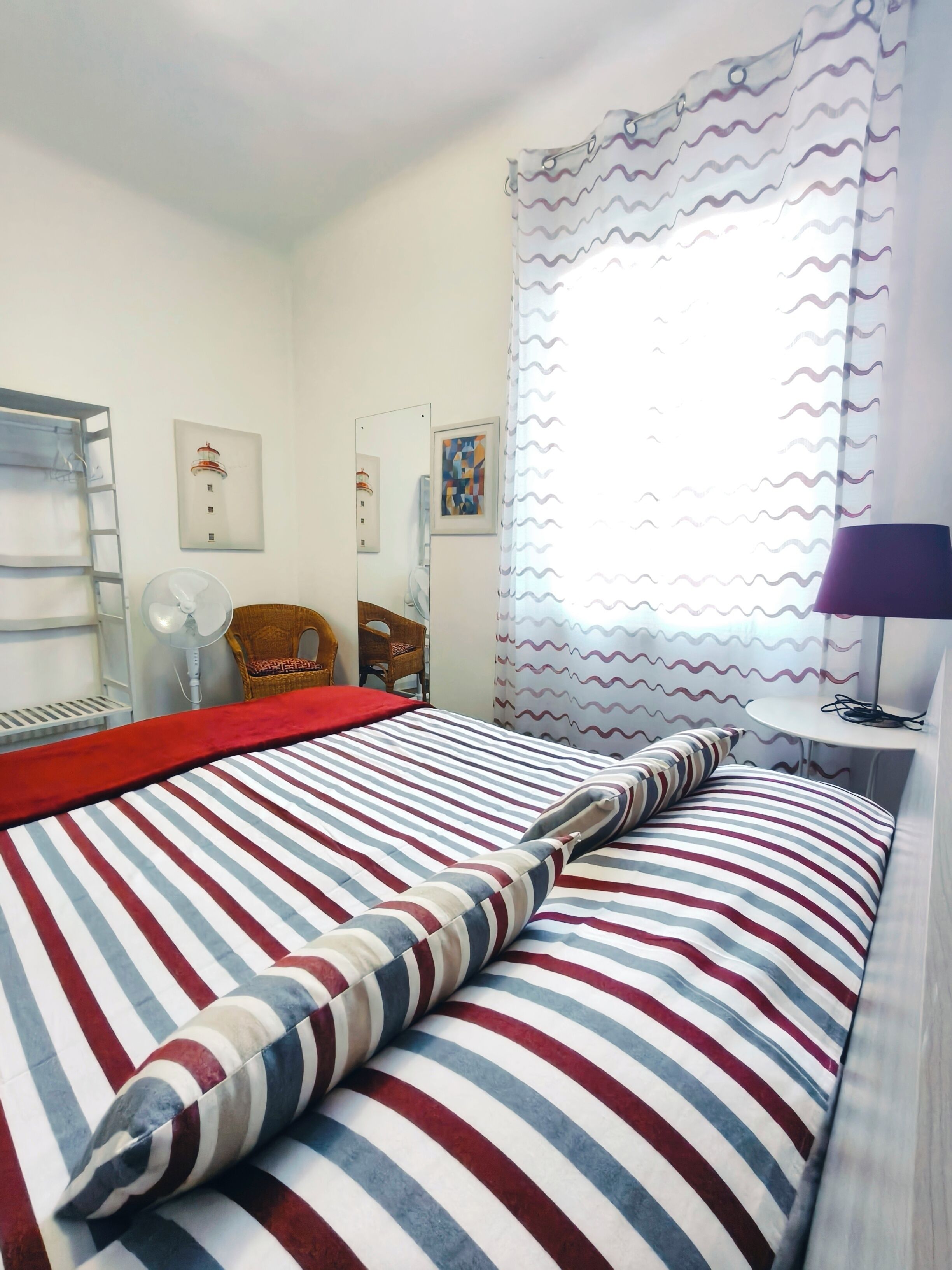 A hotel room with a bed, a chair, a table, a lamp, a window with curtains, and a wall-mounted fan.