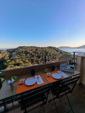 Uma varanda com uma mesa posta para dois, com vista para um morro arborizado e um corpo de água.
