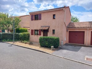Stucco building with a terracotta roof, red garage door, and two windows with shutters, set amidst a well-maintained hedge and a quiet street.