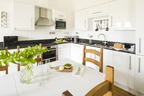 Une cuisine moderne avec des meubles blancs, un plan de travail noir, des appareils en acier inoxydable et une table à manger agrémentée de verres et d’une plante.
