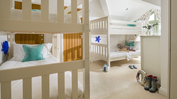 Quarto de criança com beliches, uma mesinha e uma janela com cortinas.
