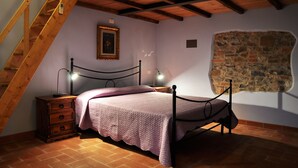 A bedroom with a wooden staircase, a stone wall, a bed with a headboard, a nightstand, and a lamp.