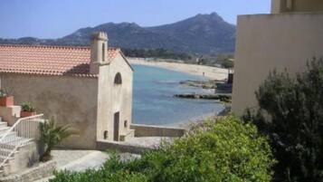 Une vue côtière avec un bâtiment doté d’un toit recouvert de tuiles rouges et d’une petite tour, surplombant une plage et des montagnes.