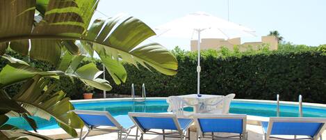 A pool area with lounge chairs and a table, surrounded by greenery.