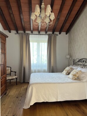 Une chambre avec un plancher en bois, un lit avec une literie blanche, un lustre, une fenêtre avec des rideaux et une chaise.