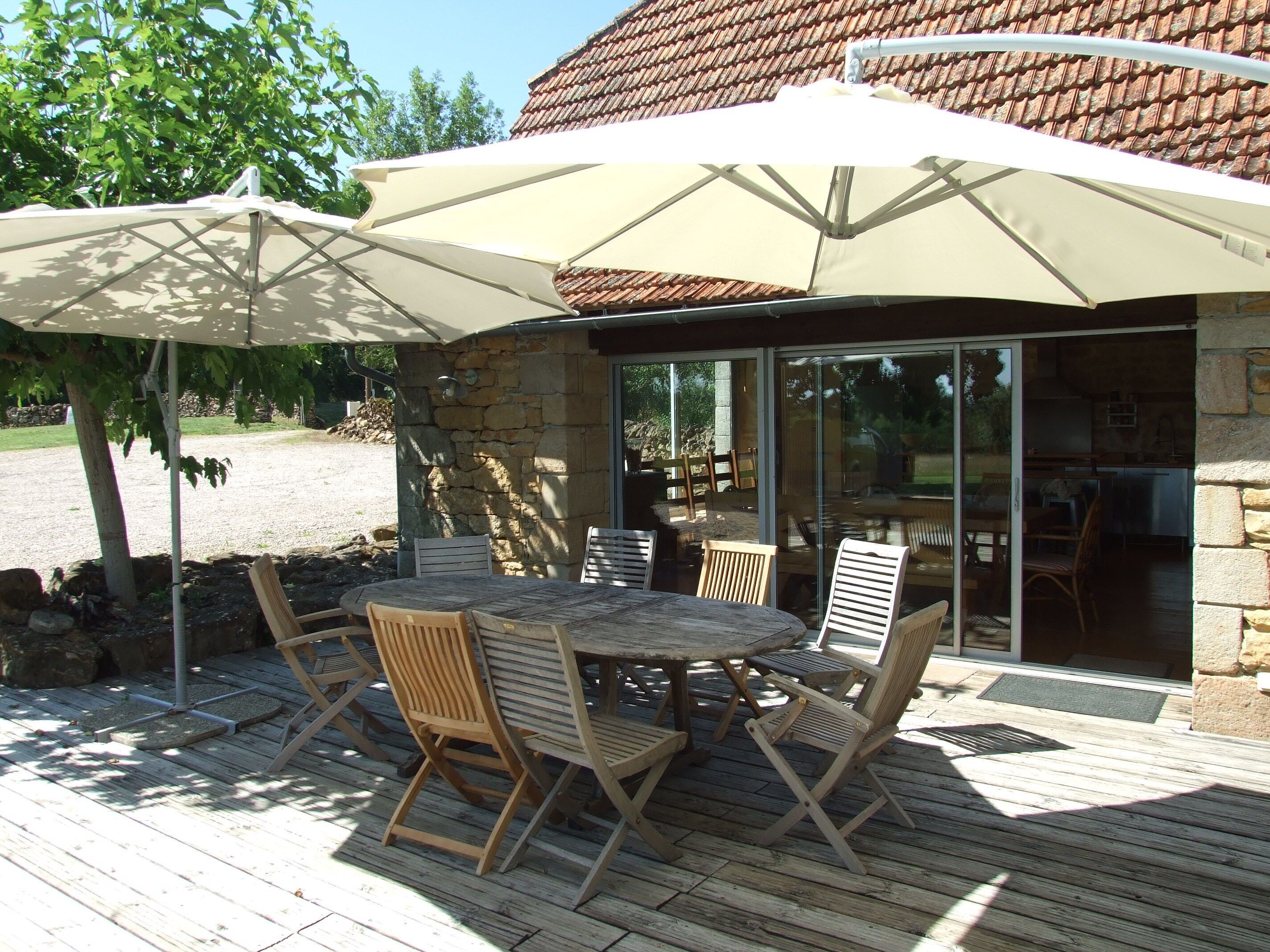 An outdoor patio with a round table and chairs, a large umbrella, and a stone building with a tiled roof in the background.