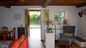 Ein gemütliches Wohnzimmer mit Holzboden, einer Couch, einem Sessel, einem kleinen Tisch mit Lampe und einem Fenster mit Blick auf einen Garten.