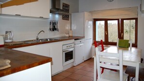 Eine Küche mit weißen Schränken, einer Holz Arbeitsplatte, einem Herd, einem Backofen und einem Spülbecken. An der Wand hängt eine Uhr und es gibt ein Fenster mit Blick auf Bäume.
