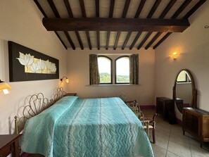 A bedroom with a bed, a window with curtains, a wall-mounted lamp, a mirror, and a decorative wall piece. - ENCHANTING FLAT  WITH PRIVATE POOL (Gualdo Cattaneo)